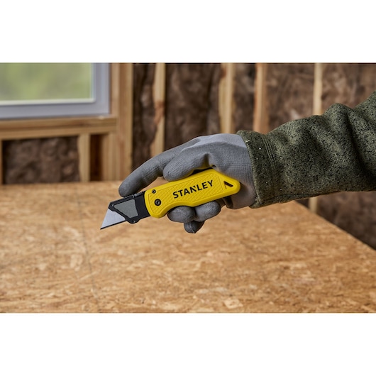 A gloved hand holding a yellow Stanley utility knife with a retractable blade, positioned above a wooden work surface in a workshop.