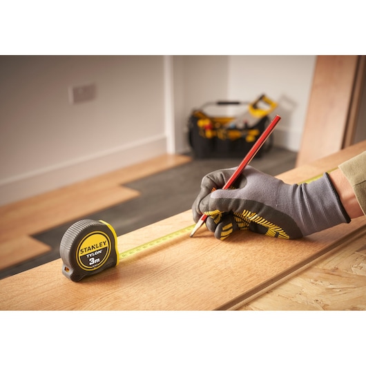 Photo of STANLEY 0-30-687 tape measure on wood, worker with gloves marking with red pencil, tool bag in background.