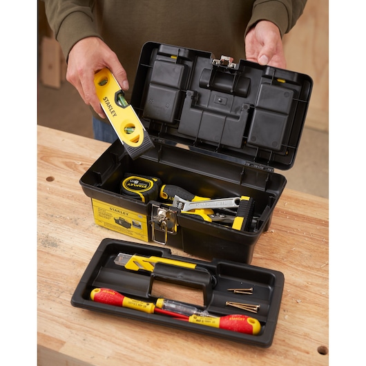 STANLEY 1-92-064 black plastic toolbox with tools and removable tray on workbench, person holding yellow level.