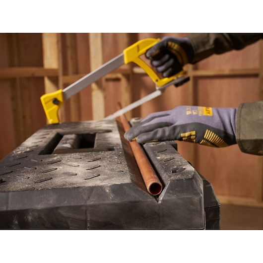 A close-up of a person wearing gloves using a yellow-handled hacksaw to cut a copper pipe on a sturdy workbench in a workshop.