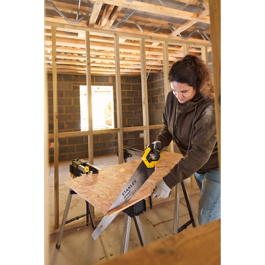 STANLEY 2-15-288 hand saw with yellow handle cutting wood board on sawhorses in construction site, side view.