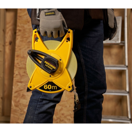 STANLEY 2-34-824 yellow 60m open reel tape measure held by worker in gloves, indoor workshop setting.
