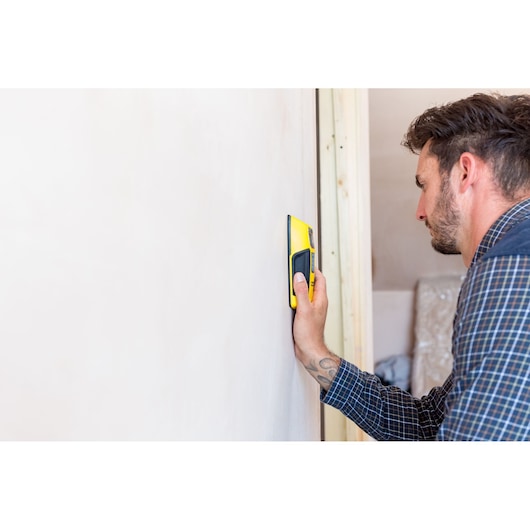 STANLEY FMHT0-77407 stud finder in hand detecting wooden stud in wall, side view, home interior.
