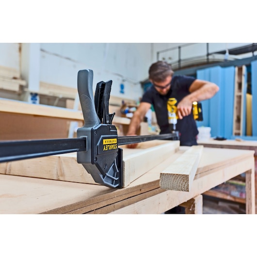 STANLEY FMHT0-83240 clamp holding wood on workbench with person working in background.