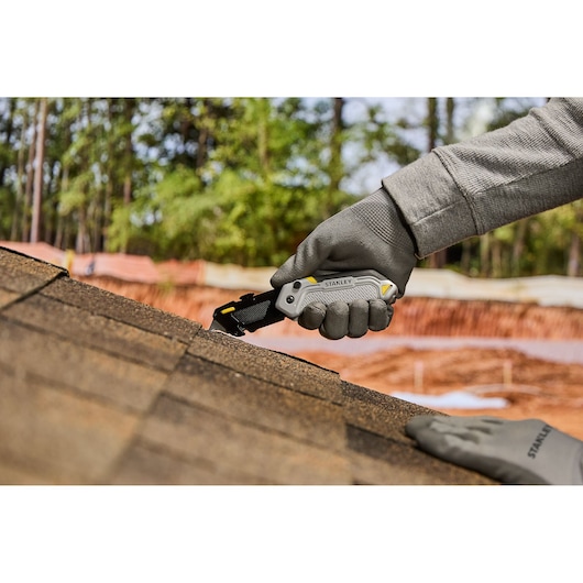 A person wearing work gloves uses a STANLEY utility knife to cut roofing shingles outdoors on a sloped roof.
