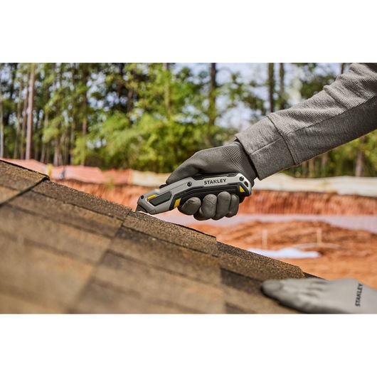 A gloved hand is using a Stanley utility knife to cut roofing shingles on a rooftop, with trees and construction materials in the background.