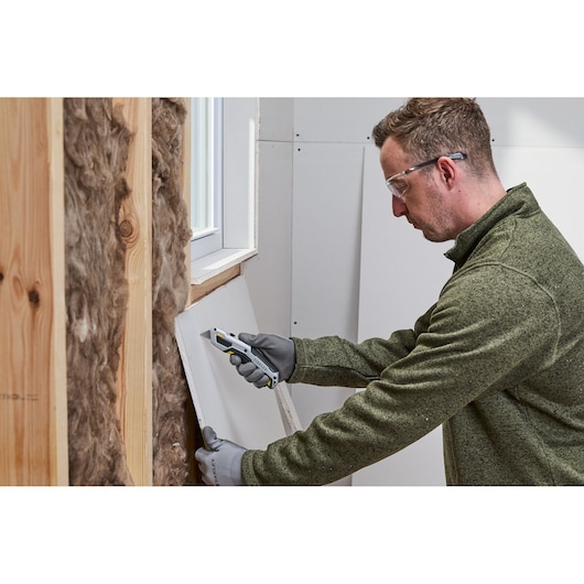 A person wearing gloves and a green sweater is using a Stanley utility knife to cut drywall near a window. The wall includes wooden studs and insulation material.