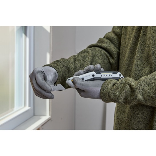 A person wearing grey gloves and a green sweater is holding a Stanley folding utility knife, replacing the blade in a well-lit indoor space near a window.