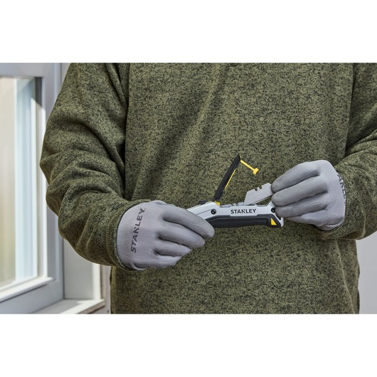 Person wearing grey Stanley gloves and a green sweater holding and demonstrating a Stanley FMHT10722-0 utility knife in front of a window.