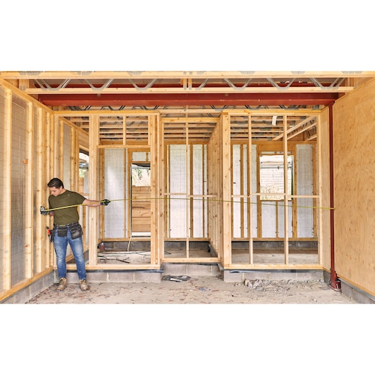 STANLEY FMHT38217-0 tape measure used by worker in wooden house construction site, measuring wall frame.