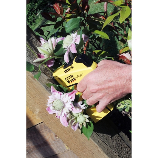 STANLEY FMHT6-70411 stapler held in hand, yellow FatMax model near pink flowers outdoors.