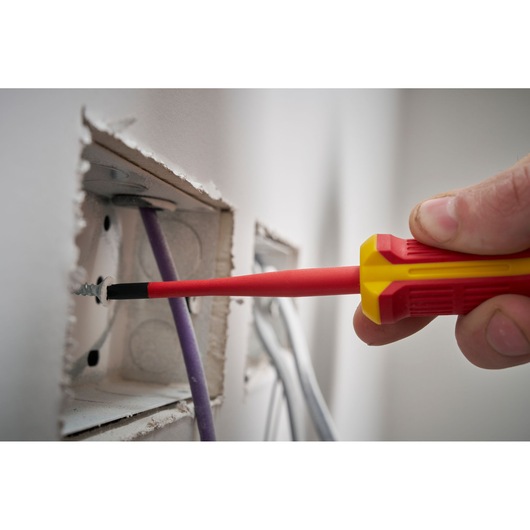 Insulated screwdriver fastening a screw inside an electrical box with visible wires in a wall opening.