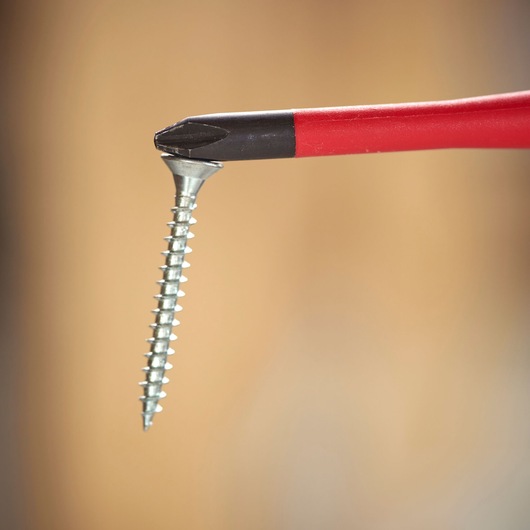 Close-up of a red-handled Phillips screwdriver tip holding a silver screw against a blurred background.