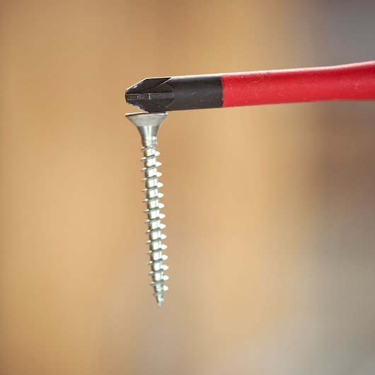 Close-up of a red-handled Phillips screwdriver tip holding a metal screw vertically against a blurred background.