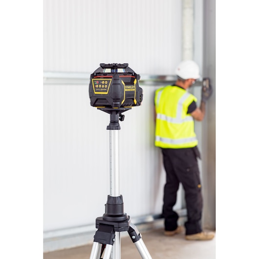 STANLEY FMHT77447-1 rotary laser on tripod, blurred worker in safety gear using tool in background.