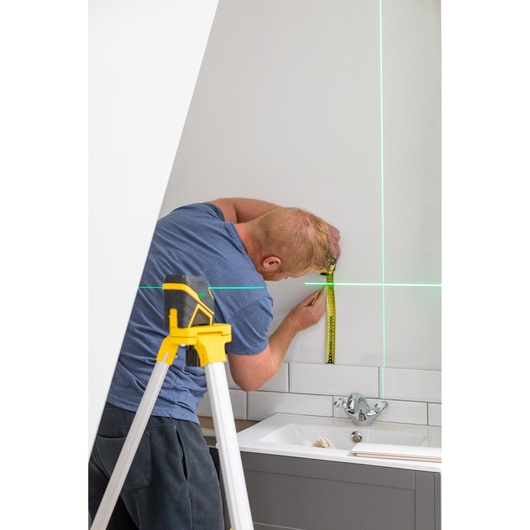 STANLEY FMHT77586-1 laser level on tripod projects green lines while man measures wall in bathroom.