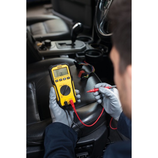 Photo of STANLEY FMHT82563-0 digital multimeter in use inside a car, with gloved hands and test leads.
