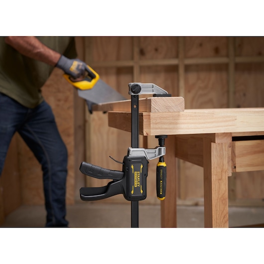 STANLEY FMHT83007-0 clamp securing wood to workbench, close-up, with person sawing in background.