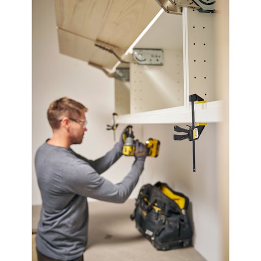 Photo of a STANLEY FMHT83270-0 bar clamp holding a white shelf, with a tool bag and person using a drill nearby.