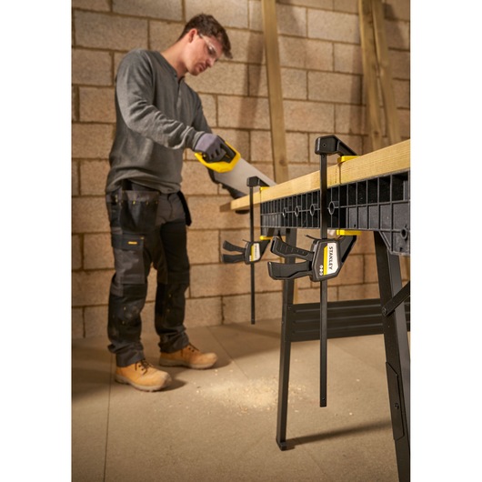 STANLEY FMHT83272-0 bar clamps securing wood on workbench, side angle, carpenter sawing in workshop.