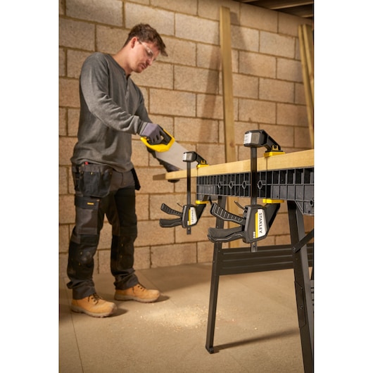 STANLEY FMHT83273-0 clamp securing wood to workbench, person sawing in workshop setting.