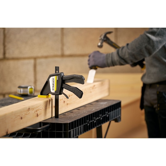 STANLEY FMHT83273-0 clamp securing wood beam on workbench with worker using hammer in background.