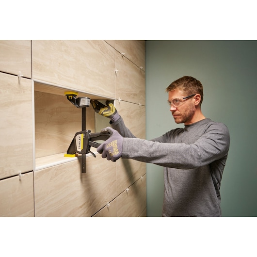 STANLEY FMHT83273-0 clamp in use on a tiled wall, person wearing gloves holding the clamp.