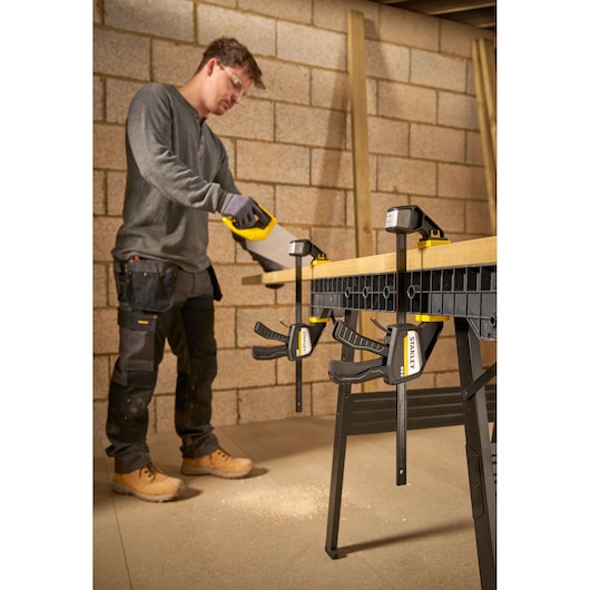 Photo of STANLEY FMHT83274-0 clamp securing wood on workbench during sawing, product in workshop setting.