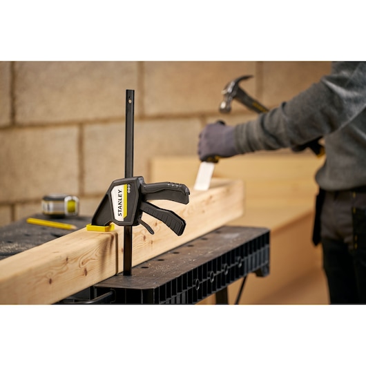 STANLEY FMHT83274-0 bar clamp secures wood beam on workbench, person using hammer in background.