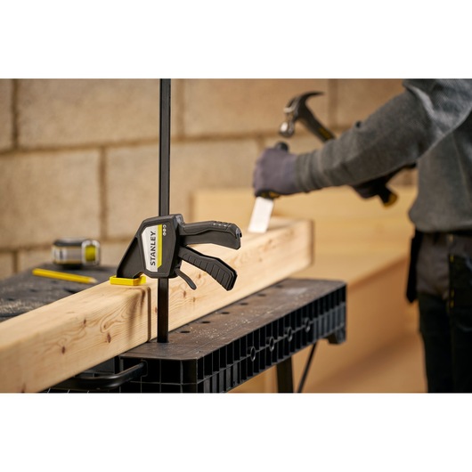 STANLEY FMHT83275-0 bar clamp securing wood beam on workbench, person using hammer in background.