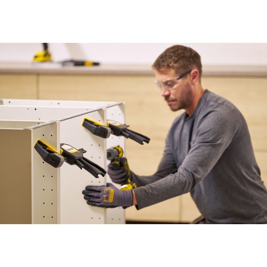 STANLEY FMHT83278-0 bar clamps holding white furniture panels, person assembling cabinet in workshop.