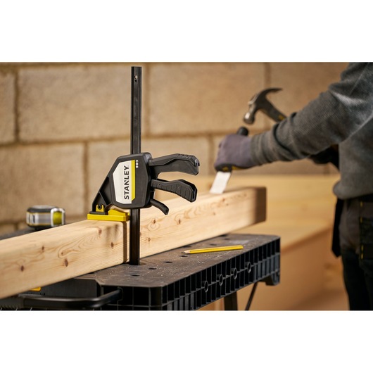 STANLEY FMHT83279-0 bar clamp securing wood on workbench, worker with hammer in background, workshop setting.