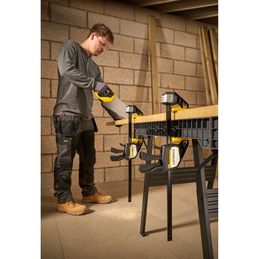 STANLEY FMHT83280-0 clamp securing wood to workbench, person sawing in workshop setting.