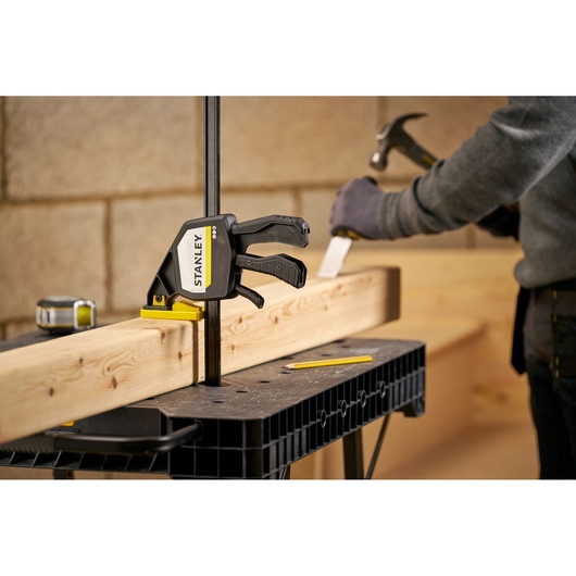 Photo of STANLEY FMHT83280-0 trigger clamp securing wood on workbench with person working in background.