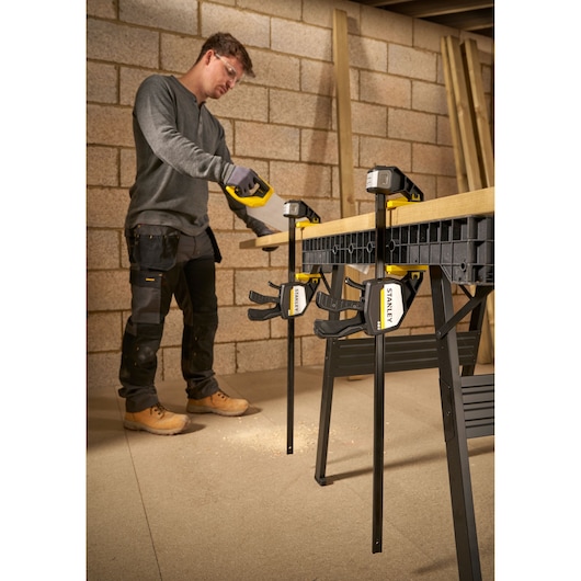 STANLEY FMHT83281-0 clamp securing wood on workbench with person sawing, workshop setting.