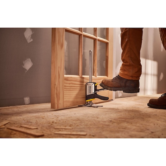 STANLEY FMHT83550-1 door installation clamp in use on a wooden door, worker using foot to operate tool.