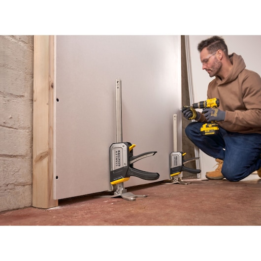 Photo of STANLEY FMHT83550-1 clamp securing drywall sheet to wall, used in construction setting.