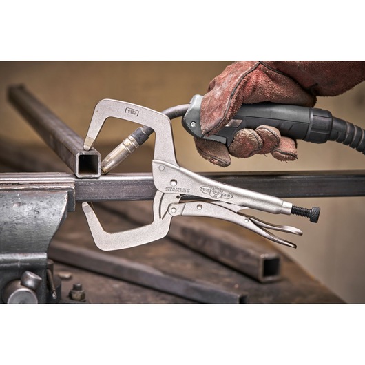 STANLEY locking C-clamp holding metal for welding, with a gloved hand using a tool on a metal workbench.