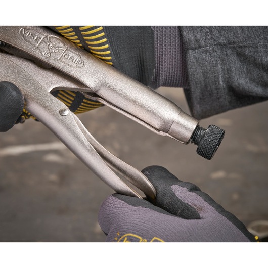 Gloved hands adjust a locking plier tool with a textured screw knob in a workshop setting.