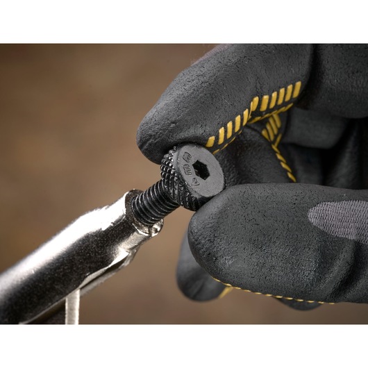 Gloved hand holding a textured screw with a wrench against a brown blurred background.