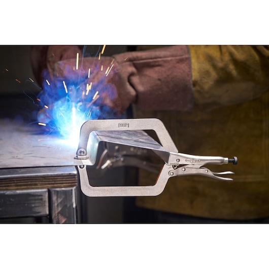 Stanley C-clamp locking pliers hold metal during welding, sparks flying, worker wears protective clothing.