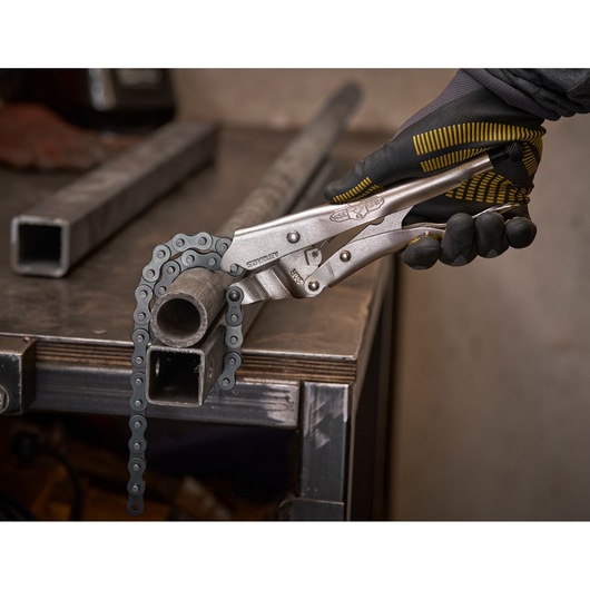 A gloved hand uses a Stanley chain locking plier to grip a round metal pipe on a workbench.