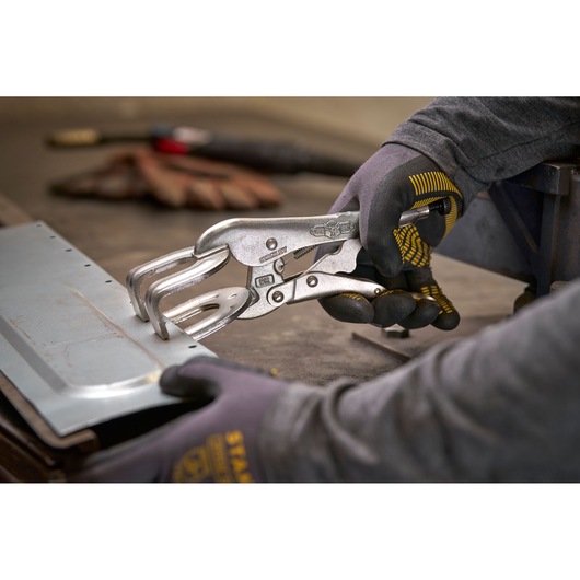 Person uses STANLEY locking pliers to hold a metal sheet on a workbench, wearing protective gloves.