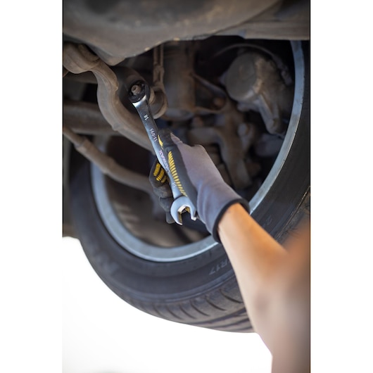 STANLEY FMMT82849-0 combination wrench used on car suspension, close-up showing gloved hand and tire.