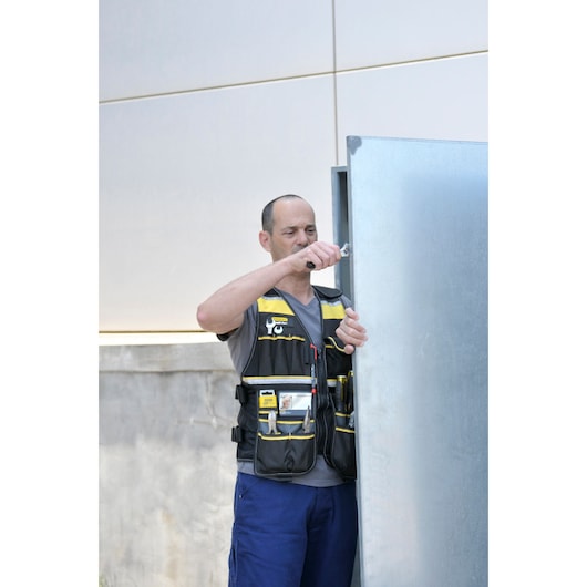 Photo of person wearing STANLEY FMST1-71181 tool vest with multiple pockets, working outdoors by a metal cabinet.
