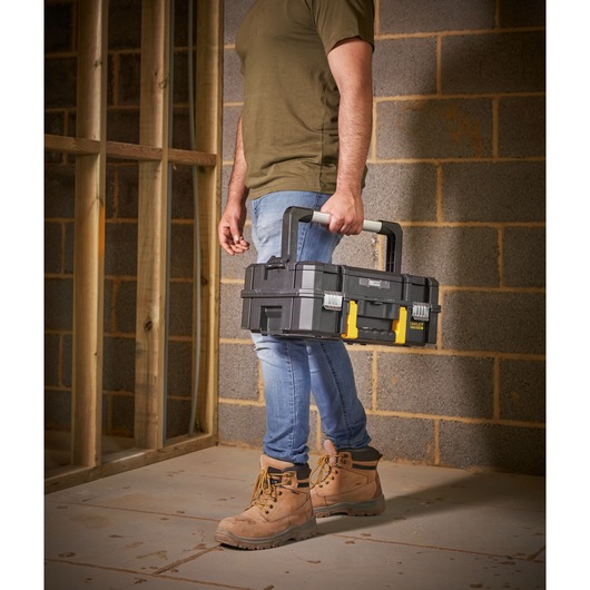 STANLEY FMST1-71967 toolbox with black and yellow latches, carried by person in work boots at construction site.
