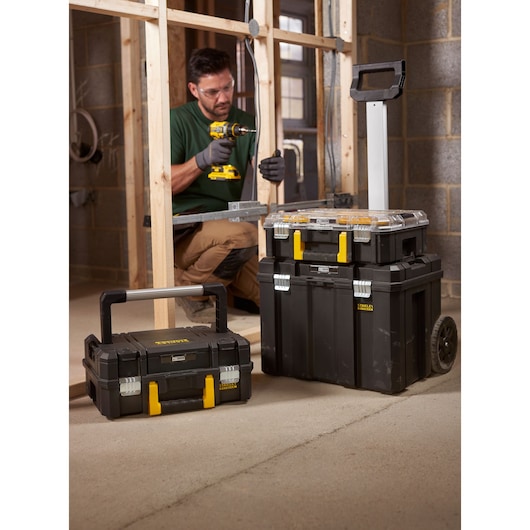 STANLEY FMST1-80103 stackable black toolboxes with yellow latches and wheeled trolley, shown on a construction site.