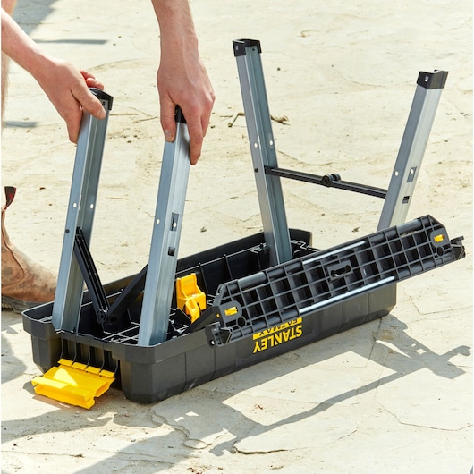 STANLEY FMST81083-1 folding workbench being assembled outdoors, showing black and yellow plastic parts and metal legs.