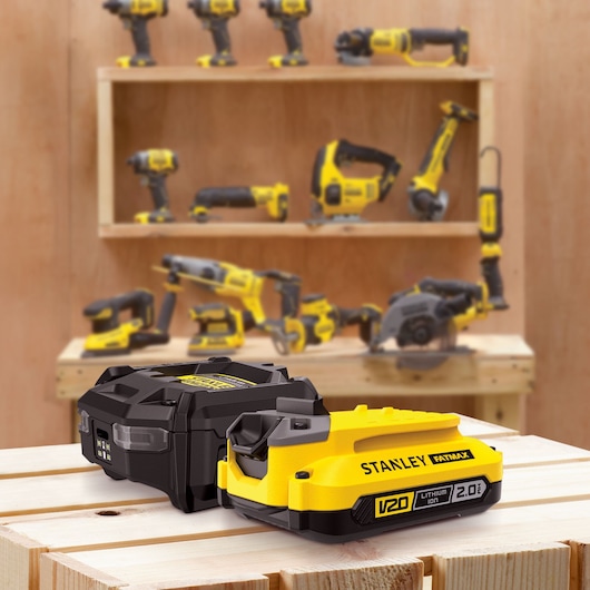 Photo of STANLEY SFMCB10D1 V20 lithium-ion battery pack on wood, with power tools blurred in background.