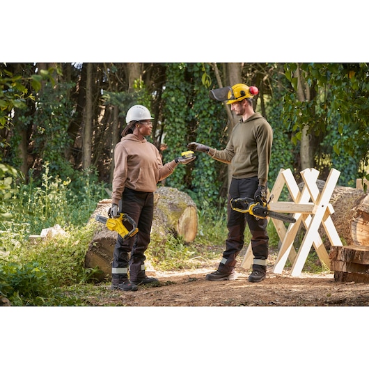 STANLEY SFMCCS730M1 chainsaw in use outdoors, two people holding chainsaws and battery in a forest setting.